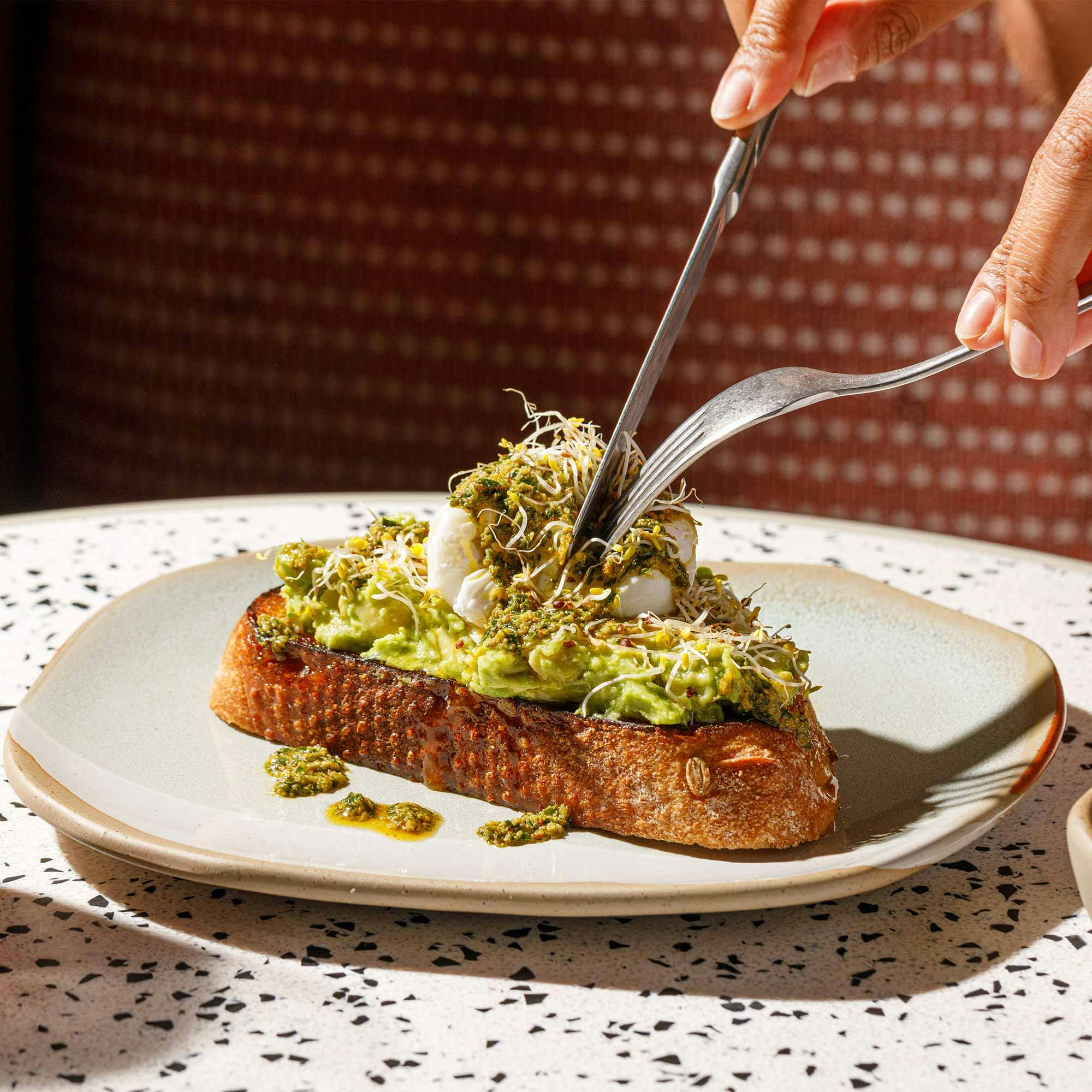 A fork punctures a herby poached egg on toast with smashed avocado, olive oil and lime.